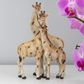 Resin giraffe family ornament showing two adult giraffes and a calf with entwined necks, in cream and brown tones with a rustic textured finish, styled with pink orchids in the background