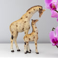 Resin giraffe mother and baby ornament showing a tall adult giraffe bending its neck down towards a small calf looking up, in cream and brown tones with a rustic finish, styled with pink orchids in the background
