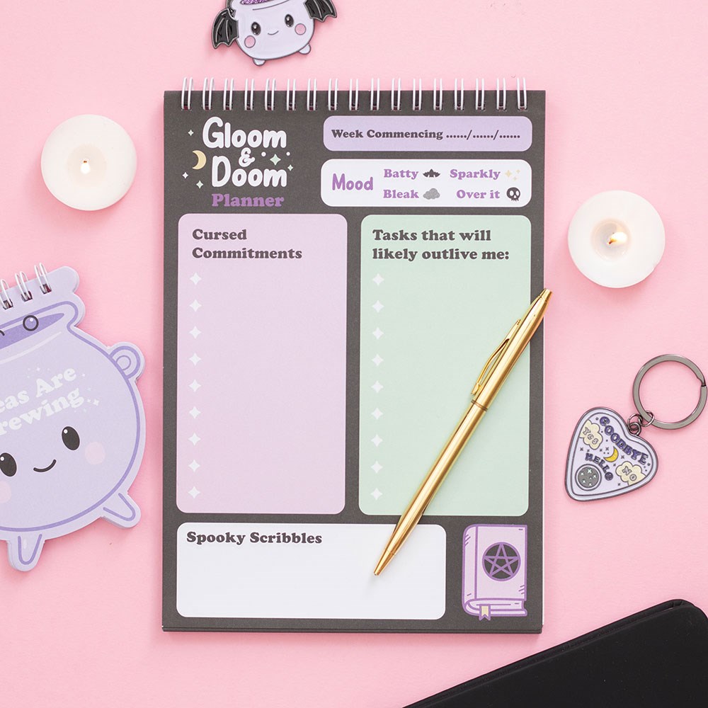 Gloom & Doom weekly desk planner open showing Cursed Commitments, mood tracker and Spooky Scribbles sections, styled with candles and a cauldron notepad on pink background