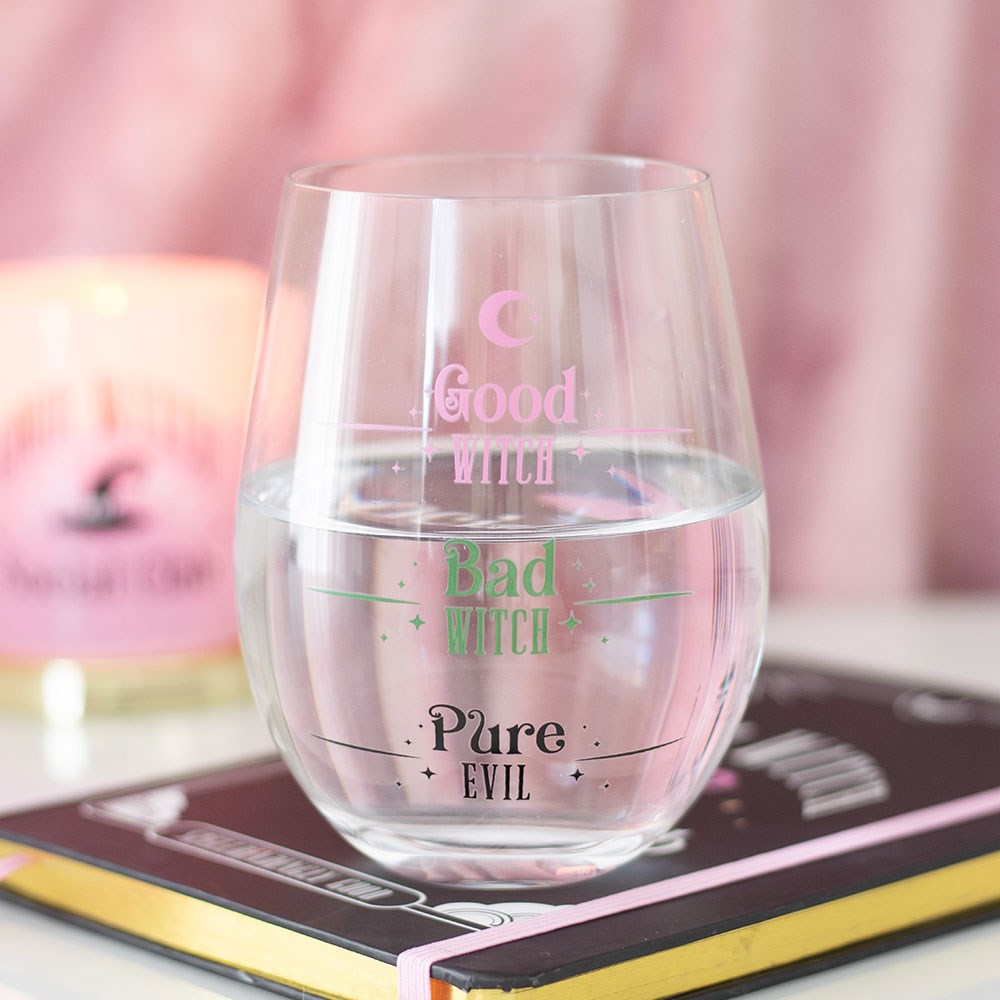 Clear stemless wine glass printed with Good Witch, Bad Witch and Pure Evil fill level markers, styled on a book with a pink candle and notebook in the background