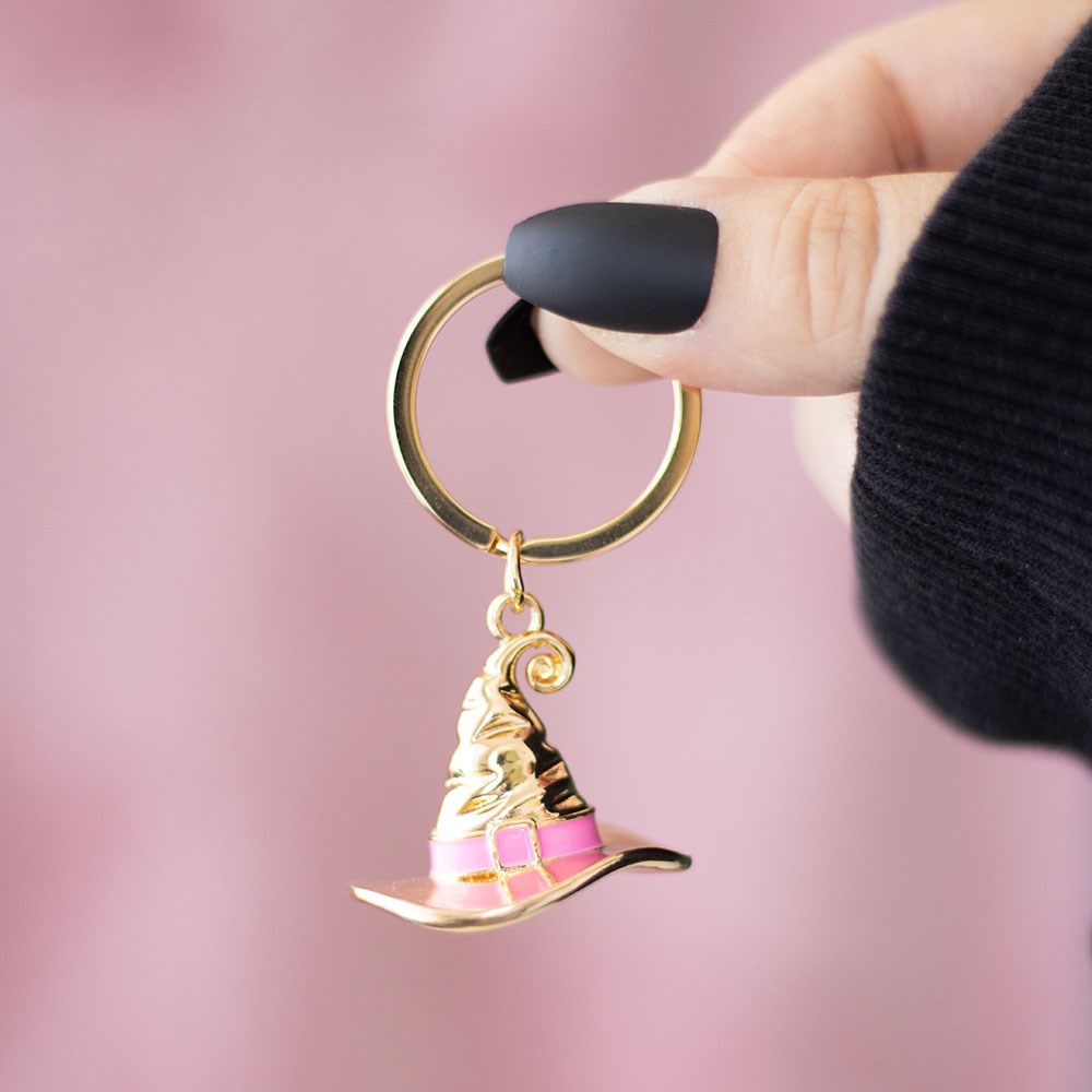 Gold-tone metal witch's hat keyring with pink enamel band detail on a gold split ring, held against a pink background