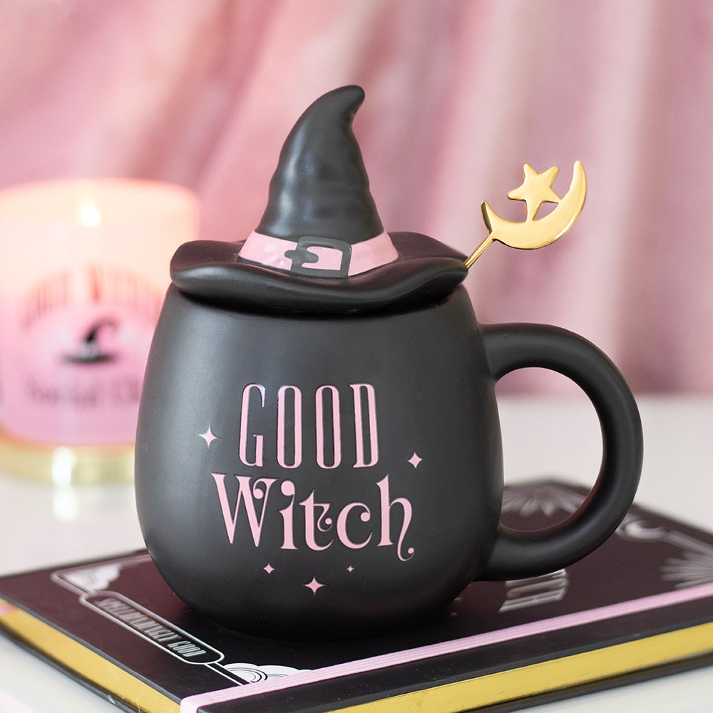 Black matte ceramic Good Witch mug with pink lettering, witch hat shaped lid and gold moon and star spoon, styled on a book with a pink candle in the background