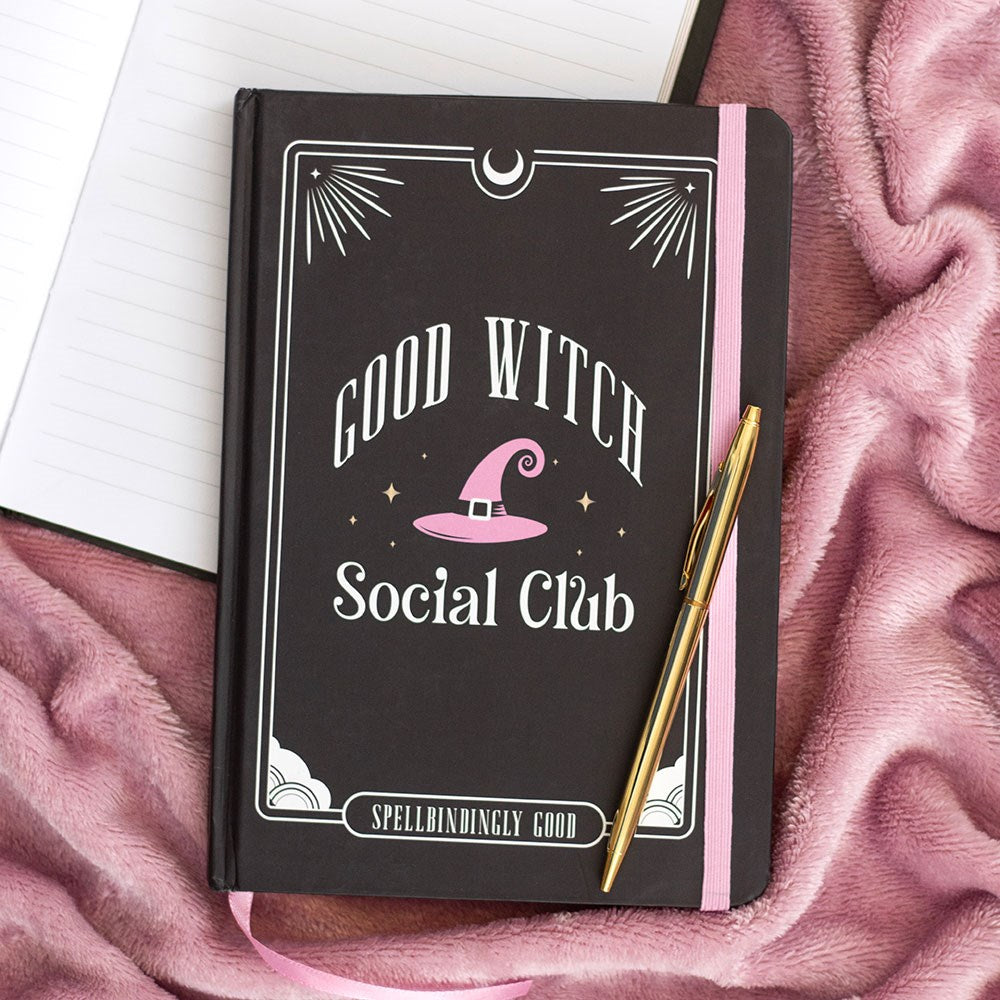 Dark hardback A5 notebook with Good Witch Social Club design, pink witch hat illustration and Spellbindingly Good text, styled on a pink fluffy blanket with a gold pen