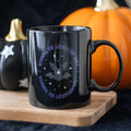 Black ceramic mug with Working The Graveyard Shift bat and star design, styled on a wooden board with Halloween pumpkins in the background