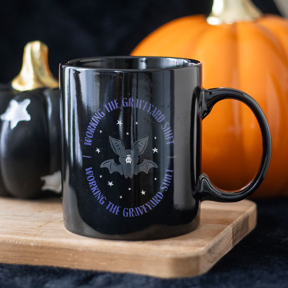 Black ceramic mug with Working The Graveyard Shift bat and star design, styled on a wooden board with Halloween pumpkins in the background