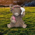 Highland cow resin figurine holding a white hairdryer and pink brush with pink hair rollers, set against a Scottish highland landscape