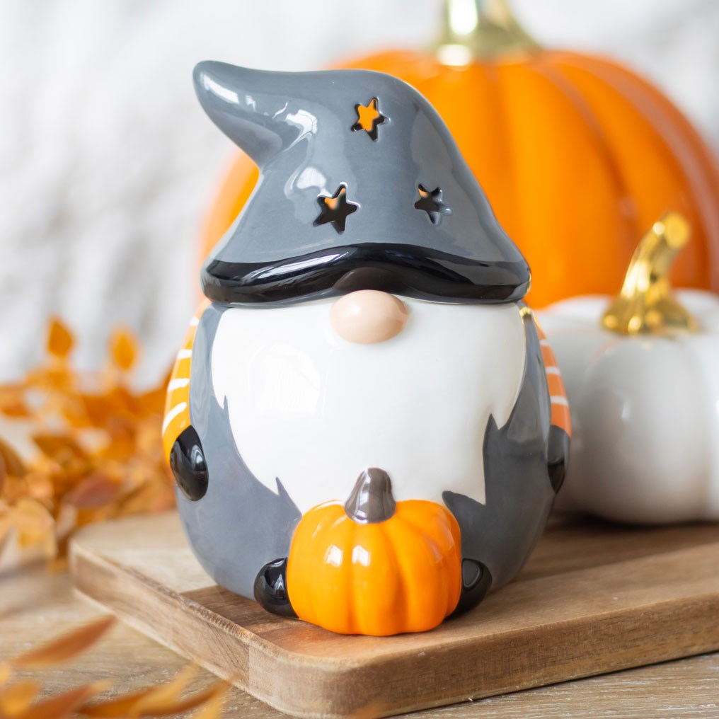 Grey ceramic Halloween gonk oil burner with star-cut witch hat, white beard, orange and black striped scarf, and orange pumpkin, styled with autumn pumpkins and leaves on a wooden board