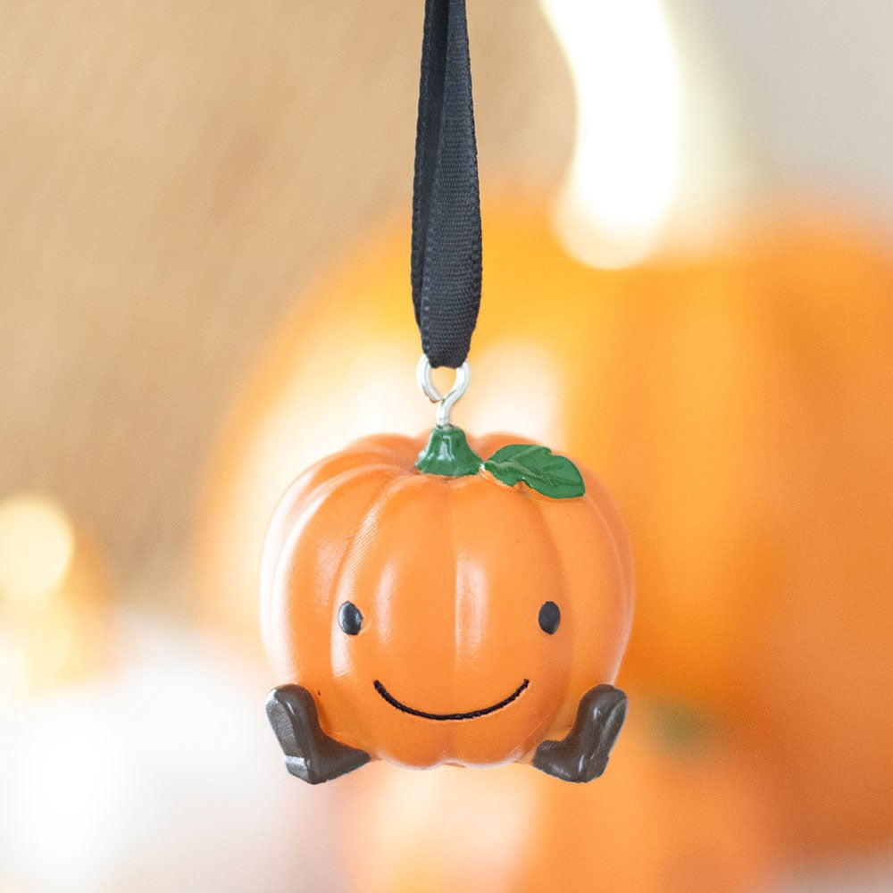 Cute orange resin smiling pumpkin hanging decoration with little dark legs and green leaf detail suspended on a black ribbon against a warm bokeh background