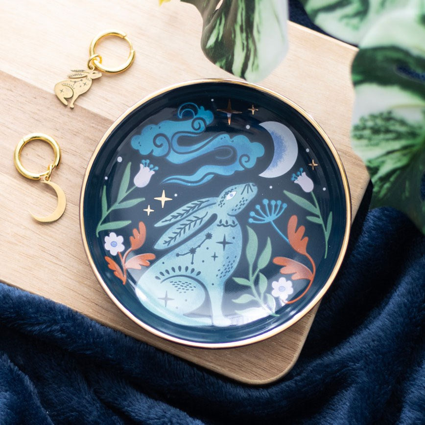 Round dark ceramic trinket dish with a gold rim featuring a mystical teal hare surrounded by orange and green botanicals, white flowers, stars, and a crescent moon, styled on a wooden board with gold hoop earrings and blue velvet fabric