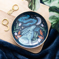 Round dark ceramic trinket dish with a gold rim featuring a mystical teal hare surrounded by orange and green botanicals, white flowers, stars, and a crescent moon, styled on a wooden board with gold hoop earrings and blue velvet fabric