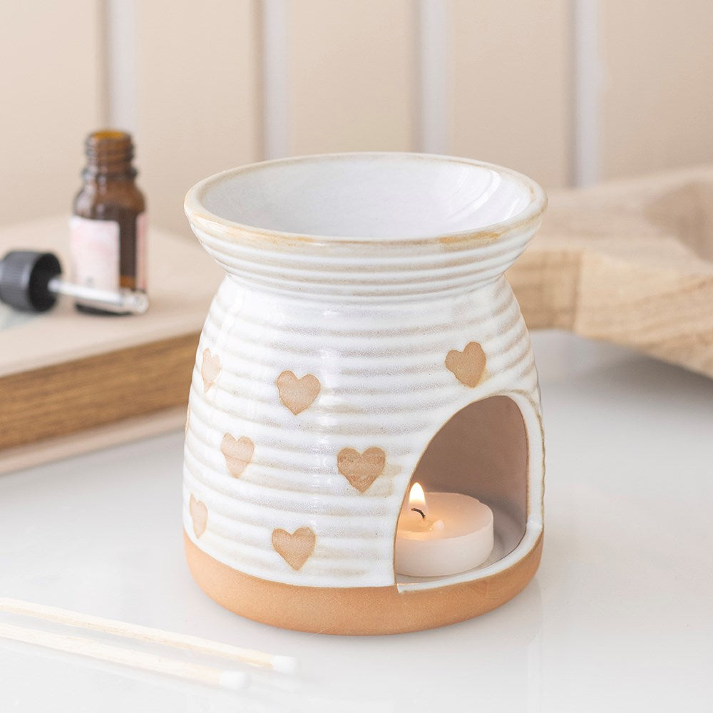 White ceramic oil burner and wax warmer with all-over heart cut-out print, ribbed texture and natural clay base, lit tealight visible inside, styled with essential oil bottle on white surface