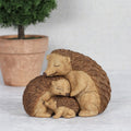 Resin hedgehog family ornament showing three hedgehogs of different sizes huddled together with eyes closed, in warm brown and cream tones, styled with a terracotta plant pot in the background