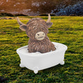 Highland cow resin figurine sitting in a white claw-foot bathtub with bubbles, set against a Scottish highland landscape