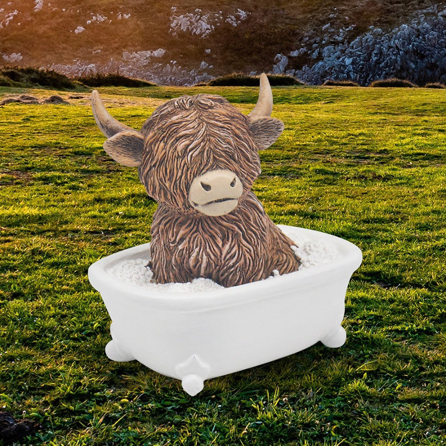 Highland cow resin figurine sitting in a white claw-foot bathtub with bubbles, set against a Scottish highland landscape