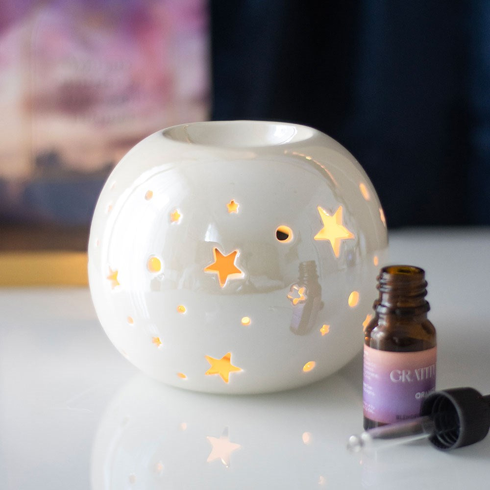 White ceramic iridescent round oil burner with star cut-outs, warm tealight glow, styled with an essential oil bottle on a white surface