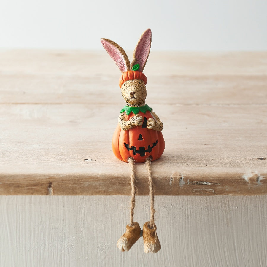 Resin rabbit figurine wearing a jack-o-lantern pumpkin costume with dangling rope legs, sitting on the edge of a wooden shelf