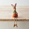 Resin rabbit figurine wearing a jack-o-lantern pumpkin costume with dangling rope legs, sitting on the edge of a wooden shelf