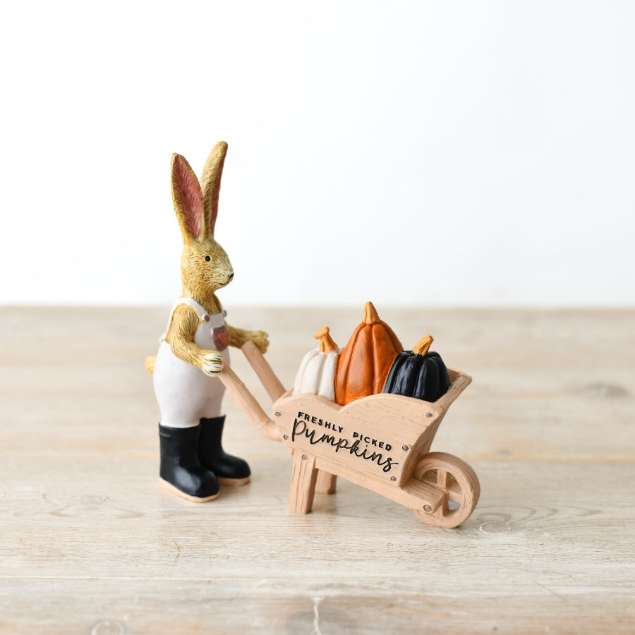 Resin rabbit figurine in white dungarees and black boots, pushing a wooden wheelbarrow filled with orange, white and black pumpkins, on a wooden surface

