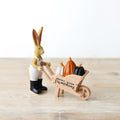 Resin rabbit figurine in white dungarees and black boots, pushing a wooden wheelbarrow filled with orange, white and black pumpkins, on a wooden surface

