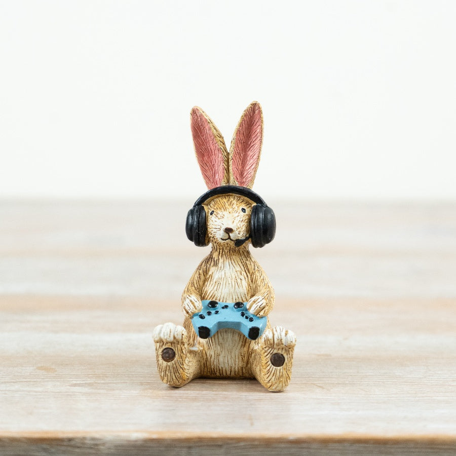 Resin rabbit figurine wearing black gaming headphones and holding a blue game controller, seated on a wooden surface against a neutral background