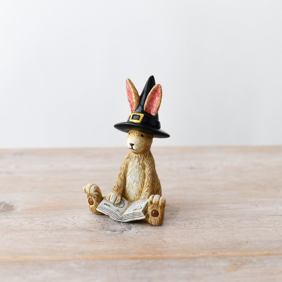 Resin rabbit figurine wearing a black witch's hat, sitting and reading a spell book, on a wooden surface

