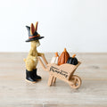 Resin rabbit figurine wearing a black witch's hat and black boots, pushing a wooden wheelbarrow filled with orange, white and black pumpkins, on a wooden surface

