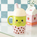 Green and pink kawaii ceramic bubble tea mug with a cream dome lid and pink straw, featuring a smiley face and brown polka dot boba design, styled on a white surface with a blue checkered background