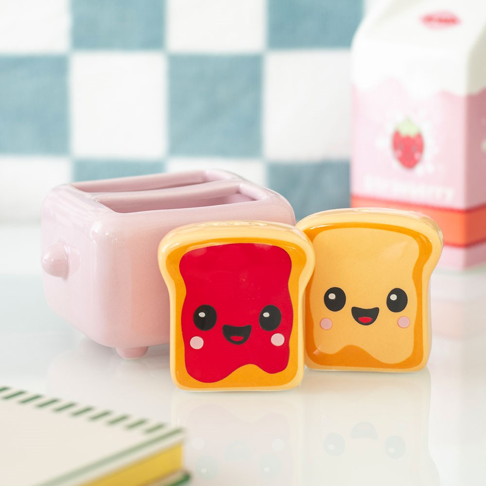 Kawaii ceramic salt and pepper pots shaped like toast slices with jam and butter smiley faces, displayed in front of a pink ceramic toaster holder on a white surface