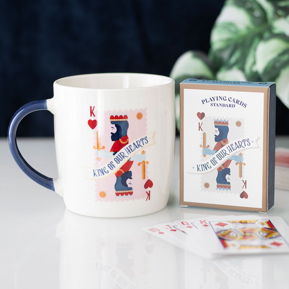 White ceramic mug with navy handle featuring King of Our Hearts playing card design, displayed next to a matching playing card box and fanned cards on a white surface