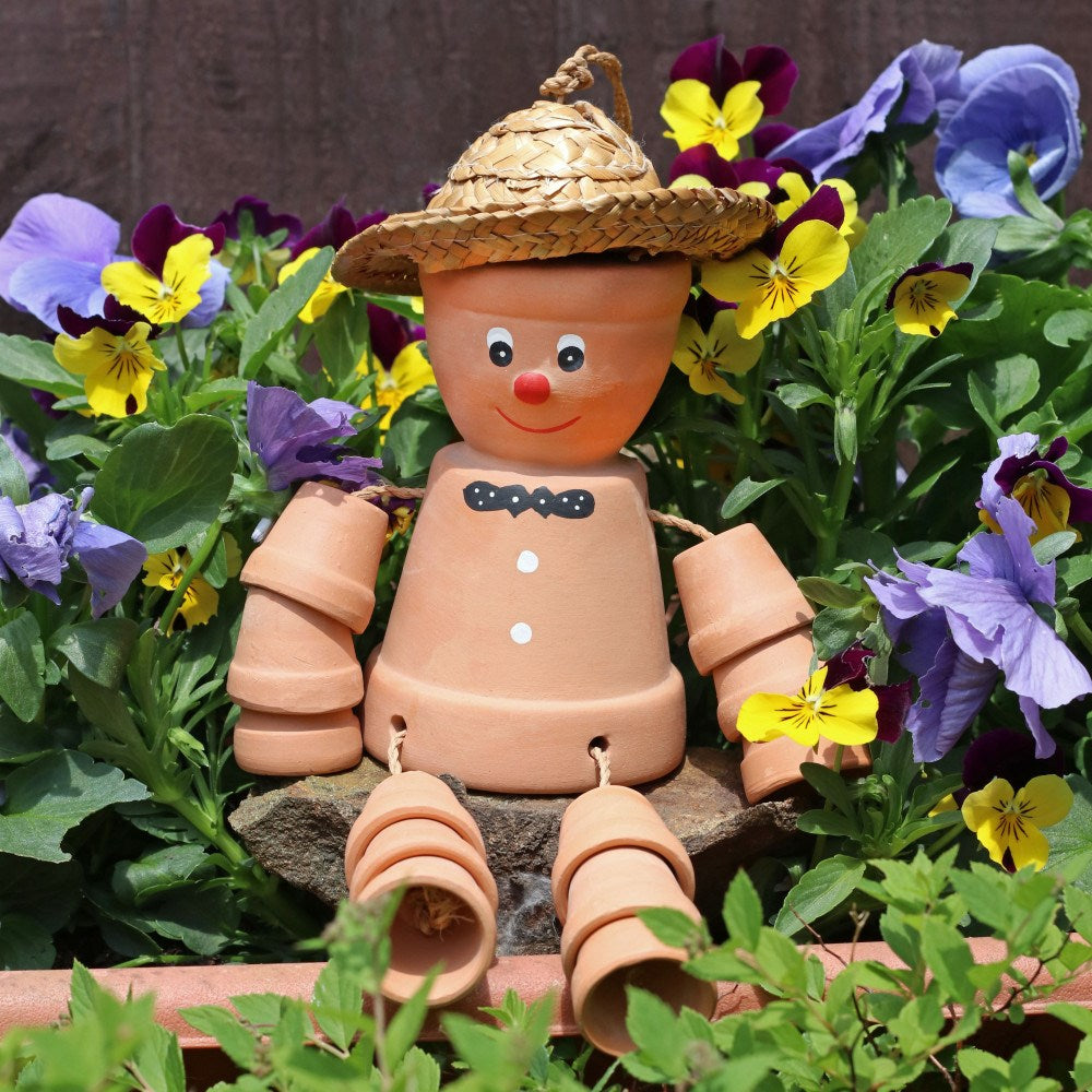 Large terracotta plant pot man ornament with movable arms and legs, wearing a straw hat and displayed among colourful garden flowers.