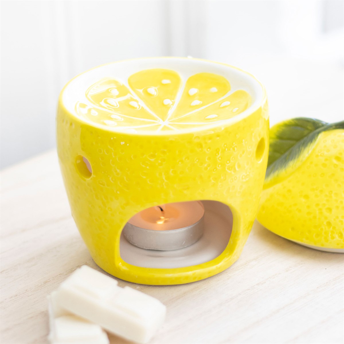 Yellow lemon shaped ceramic oil burner with citrus slice top and lit tealight, styled on a wooden board with wax melts and a fresh lemon