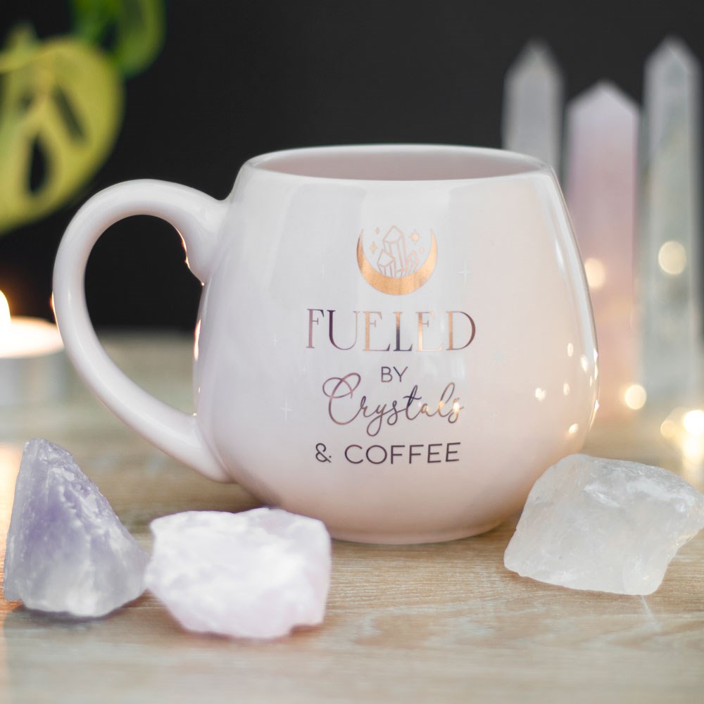 Pale pink rounded ceramic mug featuring the phrase “Fueled By Crystals & Coffee” with crystal, crescent moon and star illustrations.