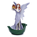 Resin fairy figurine wearing a lilac gown with large delicate wings, holding a glowing lantern aloft, standing on a green leaf base against a white background