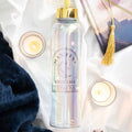 Manifest Your Life iridescent glass water bottle with gold lid and crescent moon design, styled flat lay with candles, a journal and soft white fabric
