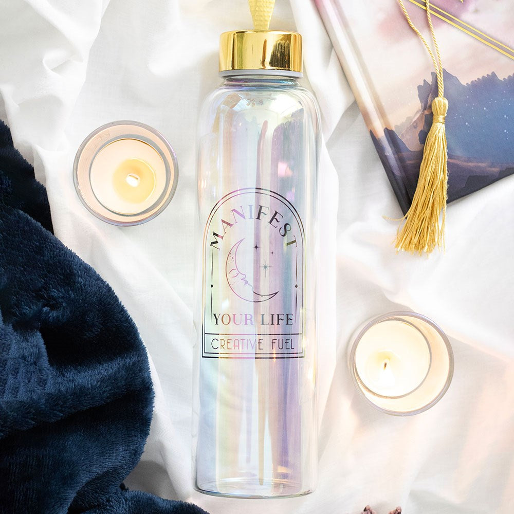 Manifest Your Life iridescent glass water bottle with gold lid and crescent moon design, styled flat lay with candles, a journal and soft white fabric