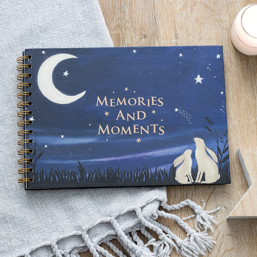 Spiral-bound baby milestone book with a dark blue cover featuring two hares gazing at a crescent moon and stars, titled "Memories and Moments", laid on a wooden surface with a grey blanket