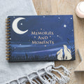 Spiral-bound baby milestone book with a dark blue cover featuring two hares gazing at a crescent moon and stars, titled "Memories and Moments", laid on a wooden surface with a grey blanket
