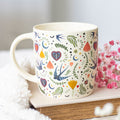 Cream ceramic mug with an all-over colourful folk art pattern featuring swallows, decorative hearts, crescent moons, and floral blooms, styled on a book with white fluffy fabric and pink gypsophila flowers