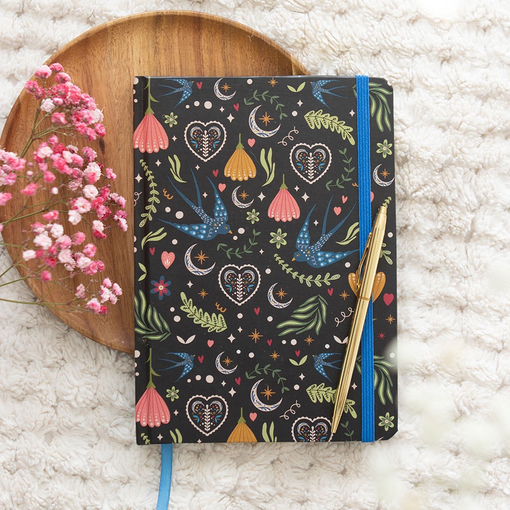 A5 lined notebook with a dark navy folk art cover featuring blue swallows, decorative hearts, crescent moons, and floral designs, with a blue elastic band and gold pen, styled on a wooden tray with pink dried flowers
