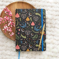 A5 lined notebook with a dark navy folk art cover featuring blue swallows, decorative hearts, crescent moons, and floral designs, with a blue elastic band and gold pen, styled on a wooden tray with pink dried flowers