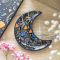 Black ceramic crescent moon shaped trinket dish with a colourful folk art pattern of blue swallows, hearts, moons, and floral designs, displayed with swallow earrings on a wooden surface with pink and white dried flowers