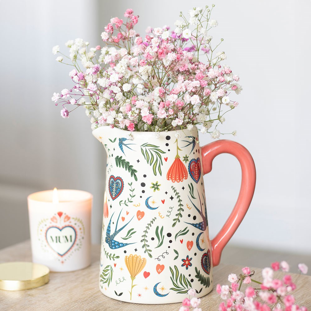 Cream ceramic flower jug with a pink heart-shaped handle and colourful folk art print of swallows, hearts, moons, and floral designs, filled with pink and white gypsophila flowers on a wooden surface