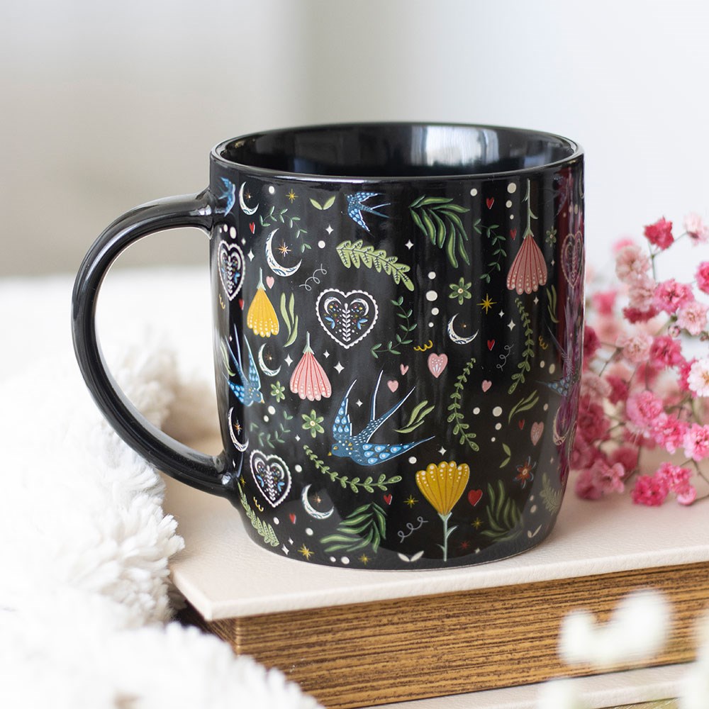 Black ceramic mug with an all-over colourful folk art pattern featuring swallows, crescent moons, decorative hearts, and floral blooms, styled on a stack of books with pink gypsophila flowers