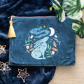 Teal velvet feel makeup bag with a mystical hare gazing at a crescent moon surrounded by green and orange botanicals and white flowers, with a gold zip and yellow tassel, styled on dark blue velvet fabric with gold star decorations