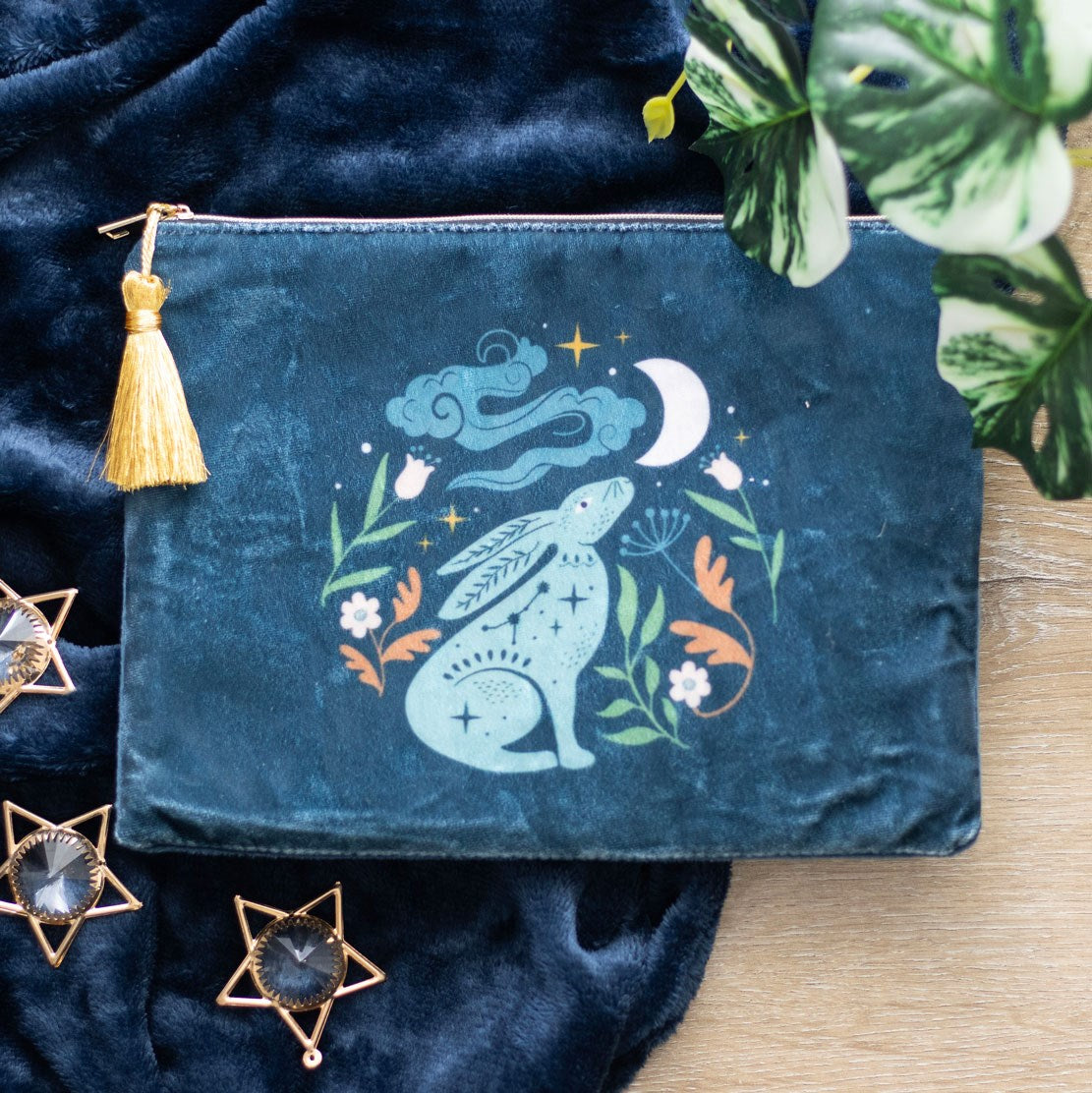 Teal velvet feel makeup bag with a mystical hare gazing at a crescent moon surrounded by green and orange botanicals and white flowers, with a gold zip and yellow tassel, styled on dark blue velvet fabric with gold star decorations