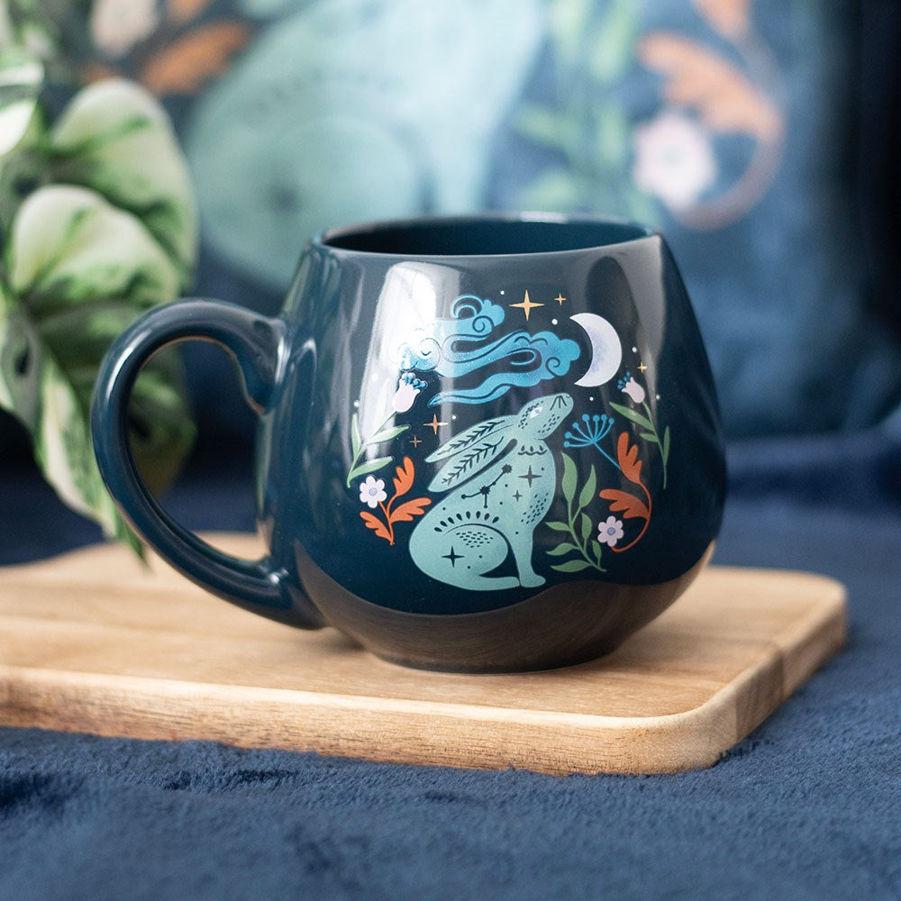 Dark teal rounded ceramic mug featuring a mystical teal hare gazing upward at a crescent moon with swirling clouds, stars, and orange and green botanical leaves, displayed on a wooden board with a blue velvet background
