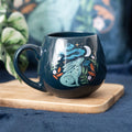 Dark teal rounded ceramic mug featuring a mystical teal hare gazing upward at a crescent moon with swirling clouds, stars, and orange and green botanical leaves, displayed on a wooden board with a blue velvet background