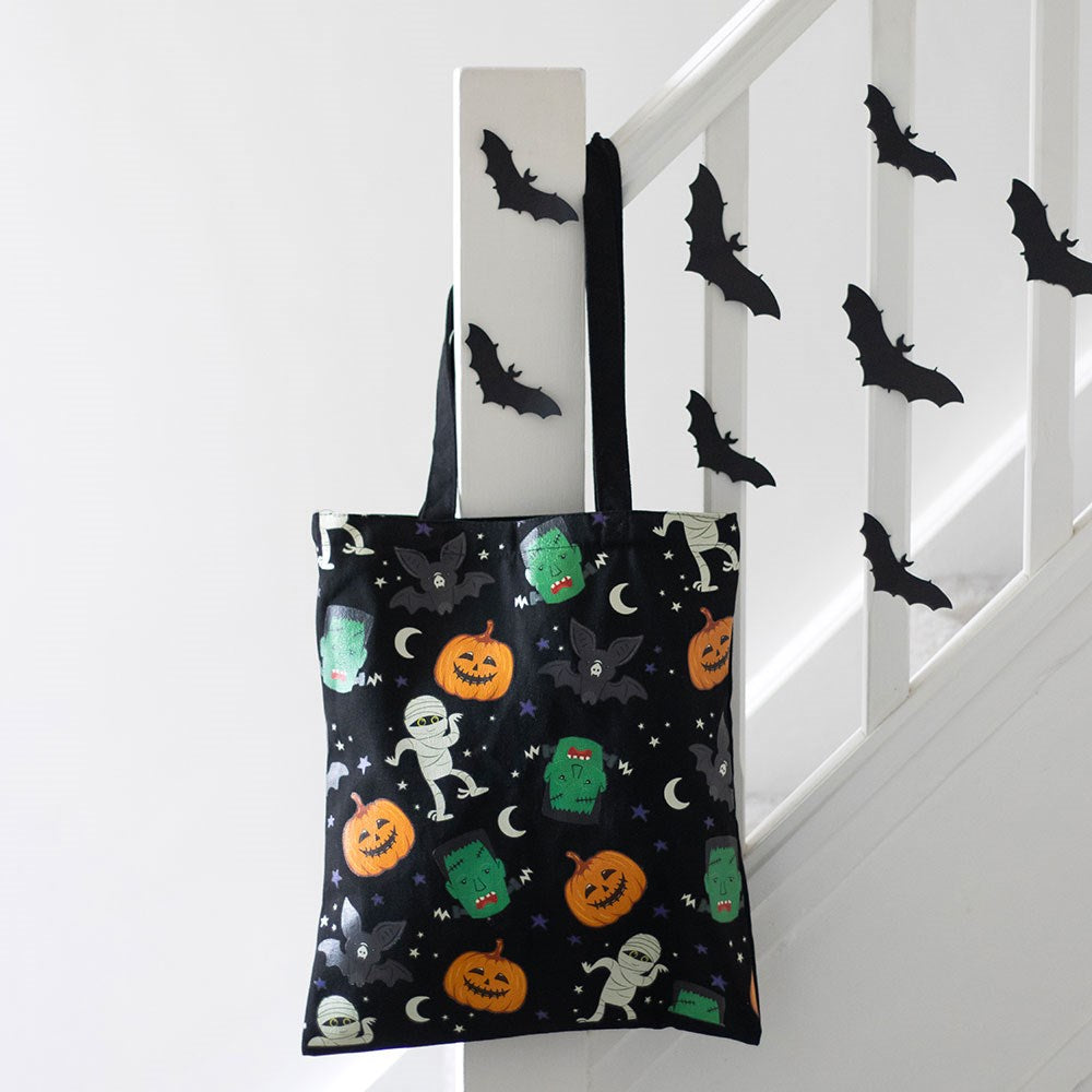 Black reusable tote bag with all-over Halloween print featuring jack-o-lanterns Frankenstein monsters mummies and bats hanging from a white staircase banister decorated with paper bats