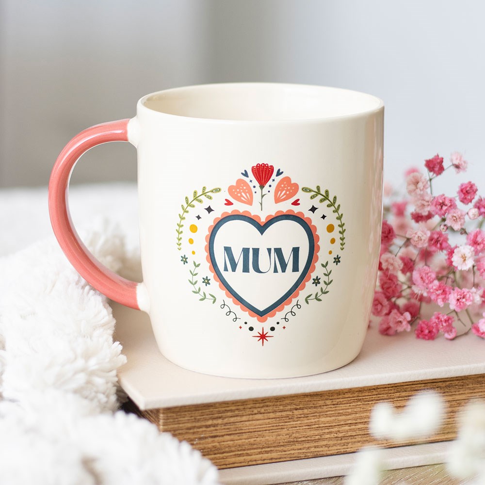 Cream ceramic mug with a pink handle featuring a colourful folk art floral heart design with "Mum" in the centre, styled on a stack of books with pink gypsophila flowers