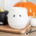 White ceramic mummy shaped oil burner with cute peeping eyes, styled on a wooden board with Halloween pumpkins in the background